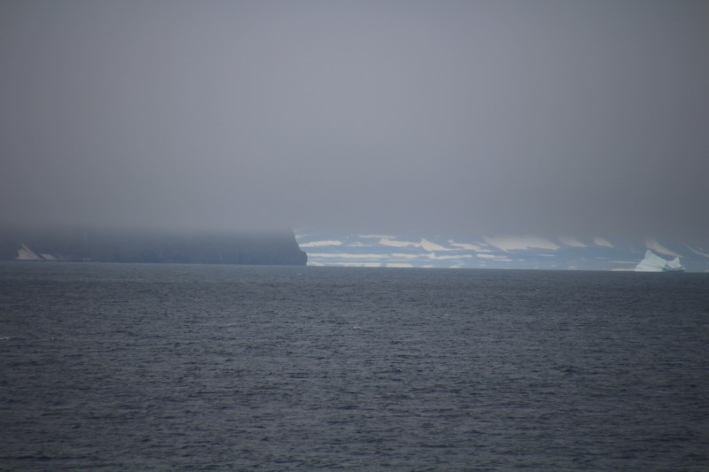 Kap Brewster in the clouds (foreground) at the southern entrance to Scoresby Sund. View is to the north-west into the entrance of the fjord. In the background the northern shores are visible behind the iceberg. [Photo by Dragonfly Leathrum]